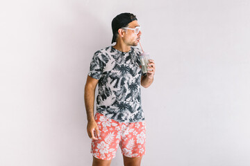 Young man with floral shirt, cap and glasses in summer drinking a cocktail with a straw, in daylight on a white wall