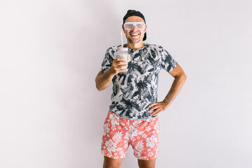 Young man with floral shirt, cap and glasses in summer drinking a cocktail with a straw, in daylight on a white wall