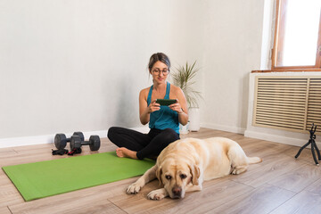 A woman in a blue T-shirt is resting after a home workout with a phone in her hands, her dog is lying nearby
