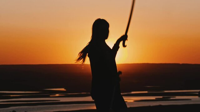 Young woman in a hoodie swinging with a blade on late orange sunset