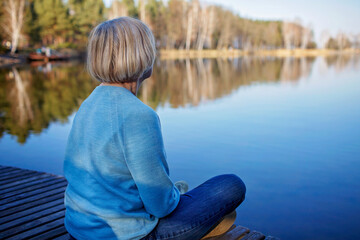 Senior woman in blue pullover sits on the shore of lake, have a rest and enjoys calm and nature, mental health, early fall, outdoor lifestyle, back view