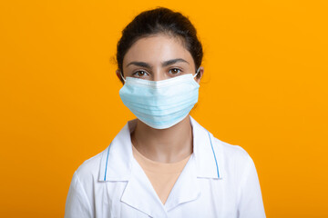 Portrait of indian doctor woman wearing white medical gown and protective surgical face mask isolated on yellow background.