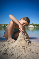 A little cute girl in a swimsuit is building a sand castle on the shore of a blue quarry lake.
