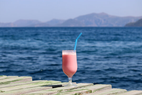 Glass With Pink Cocktail And Straw On A Wooden Pier On Sea And Mountains Background. Summer Vacation On A Beach