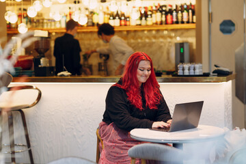 European plus size woman uses laptop while sitting on cafe indoor. Young focused red pink haired...