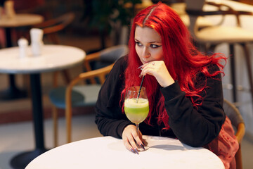 European plus size woman drink green smoothie in cafe. Young red pink haired body positive girl...
