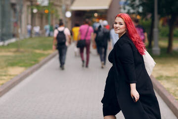 European plus size woman walking outdoor street city. Young red pink haired body positive girl.