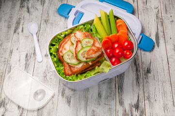 Heated lunch box for carrying and storing food. Next to it is a lid and a spoon. In the boxing sandwich and vegetables for a snack. View from above