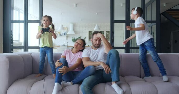 Active kids indulge jumping with digital gadgets on sofa close to their exhausted and bored parents. Cute girl plays with joystick and handsome boy listens to music on headphones. 4 K video footage