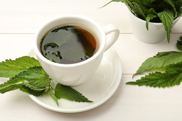 Cup of aromatic nettle tea and green leaves on white wooden table, closeup