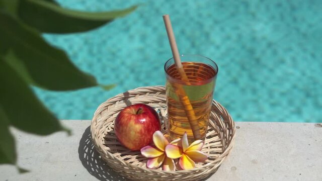 Video footage of glass with apple juice, bamboo straw, red apple, tropical flower frangipani, branch of tree with green leaves and bubbling blue swimming pool on background.
