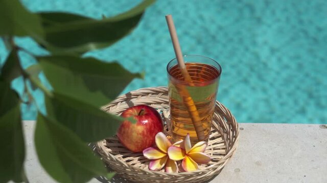 Video footage of glass with apple juice, bamboo straw, red apple, tropical flower frangipani, branch of tree with green leaves and bubbling blue swimming pool on background.
