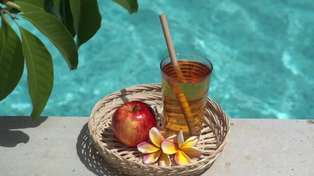Video footage of glass mason jar with apple juice, bamboo straw, red apple, tropical flower frangipani and bubbling blue swimming pool on background.