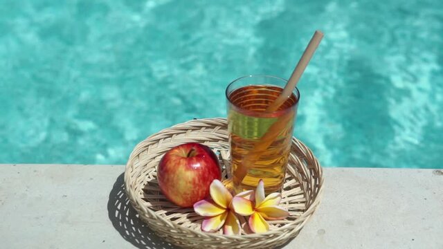 Video footage of glass with apple juice, bamboo straw, red apple, tropical flower frangipani and bubbling blue swimming pool on background.
