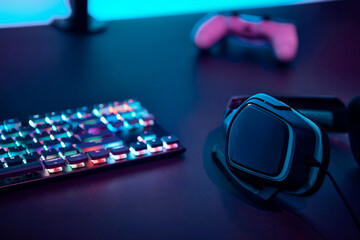 Headphones, colorful backlit gaming keyboard and gamepad laying on dark desk.