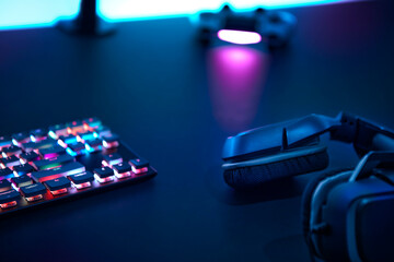 Headphones, colorful backlit gaming keyboard and gamepad laying on dark desk.