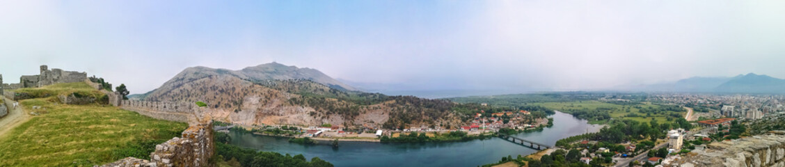 Fototapeta na wymiar Panorama of the Buna river valley - view from the wall of the Shkoder castle (Albania). Beautiful widescreen landscape with the ruins of an ancient fortress, mountains, plain, river and city