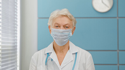 Fototapeta premium Grey haired senior woman doctor wearing surgical mask and white robe with stethoscope looks into camera