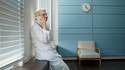Happy mature lady doctor with glasses talks on cellphone with family member during at short break...