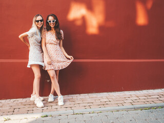 Fototapeta na wymiar Two young beautiful smiling hipster female in trendy summer dresses.Sexy carefree women posing on the street background near red wall. Positive pure models having fun at sunset, hugging
