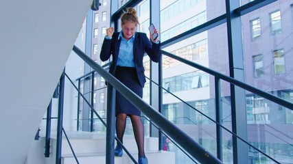 A young office woman with smartphone is descending the stairs making yes gestures. Ecstatic dance of a winner in modern building. A concept of sudden lottery or giveaway winning, or date invitation