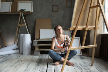 Fototapeta na wymiar A young beautiful woman artist in a dirty T-shirt sits on the wooden floor in an art studio, holds brushes in her hands and looks at an easel with a picture.