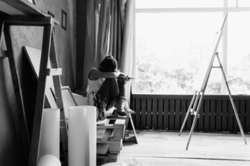 Depression in a woman artist, a young lady sits in an art studio near an easel and canvas, bowed...