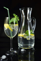 Refreshing drink with lemon, mint and ice in a jug and glass on a black background