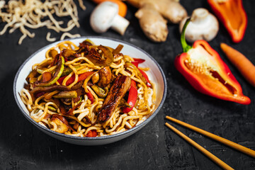 Wok with turkey meat, noodles, paprika, mushrooms and carrot served on a black background with chopsticks