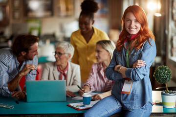 young adult redhead female posing at work in front of group of working people, smiling, looking at...