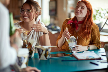 young female colleagues having a break at work eating takeaway chinese food with sticks, smiling