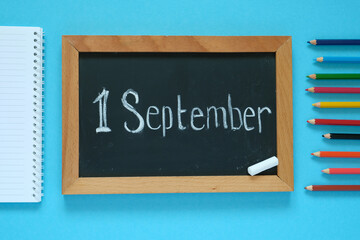 Flat lay. Blue background. In the center is a chalk board with the inscription "September 1". Nearby is a notebook and pencils. Knowledge day, school, training, business.
