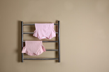 Modern heated towel rail with baby clothes on beige wall. Space for text