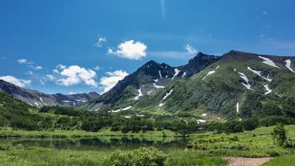 A picturesque lake in the valley, next to a mountain range. There is green vegetation on the banks, snow patches on the slopes. Reflection on the water surface. Blue sky. A summer day. Kamchatka