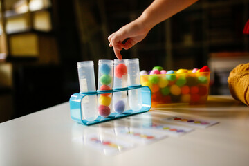 Obraz na płótnie Canvas Cute Caucasian child on the table plays with balls and test tubes, the child studies the color using the game. Sensory development and homemade lessons, dark style in the real interior, close up