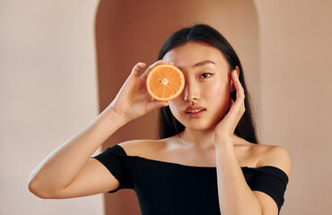 With slice of orange. Young serious asian woman standing indoors