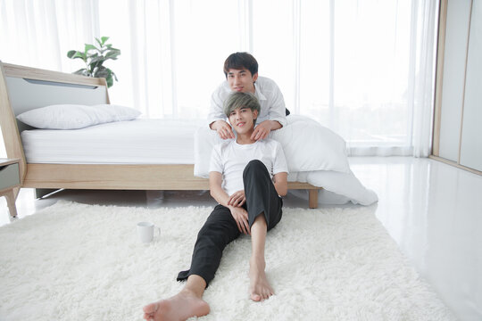 LGBT Asian gay couple in white bedroom. Young man smiling lying on bed putting his chin on boyfriend's head who sitting on floor with cup of coffee. LGBTQ in mood of love concept