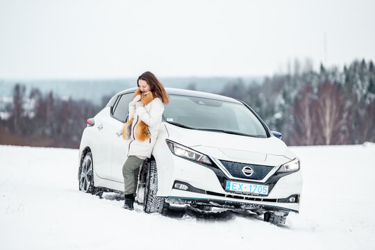 Aluksne, Latvia - 01.12.2021 Beautiful Woman Standing Near Metallic White Nissan Leaf In Winter. Compact Five-door Hatchback Electric Car Manufactured By Nissan