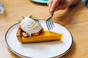 Orange carrot pie with whipped cream on top on white plate and woman hand with fork at wooden table in vegan cafe closeup