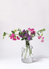 A bouquet of pink and lilac wildflowers in a small transparent vase on a white background