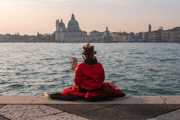 Male in a red carnival costume sitting by the water looking at the Church of San Giorgio Maggiore