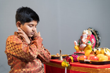 studio shot of kid by holding ears doing squats in front of lord ganesha during festival...