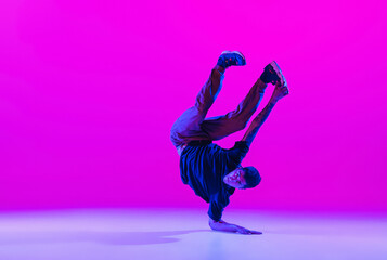 Young stylish man, hip-hop dancer dancing solo in modern clothes isolated over bright magenta background at dance hall in neon light. - obrazy, fototapety, plakaty