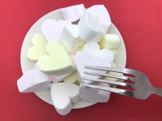 白いお皿に乗ったハート型のマシュマロとフォーク（フォーク下・赤背景）、バレンタイン・ホワイトデーイメージ