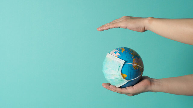 Hand is hold earth globe with face mask on mint green or Tiffany Blue background.