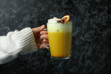 Woman in sweater holds glass of pumpkin latte on black smokey background
