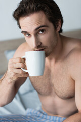 young shirtless man drinking morning coffee and looking at camera in bedroom