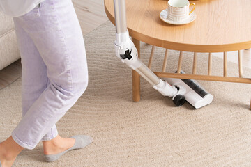 young woman vacuums carpet under table with cordless vacuum cleaner. Female legs. Close-up of...