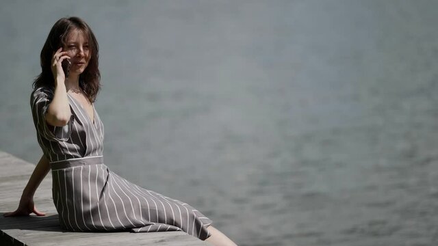 a brunette in a striped dress is sitting on a wooden deck by the river on a sunny day. she talks on the phone, rubs lower back, swings legs