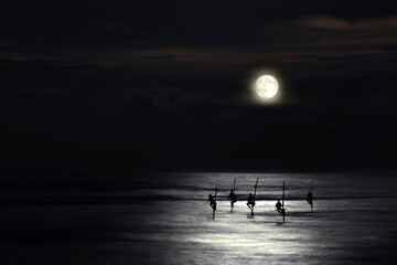 Fishermen on stilts in silhouette in the moonlight in Galle, Sri Lanka